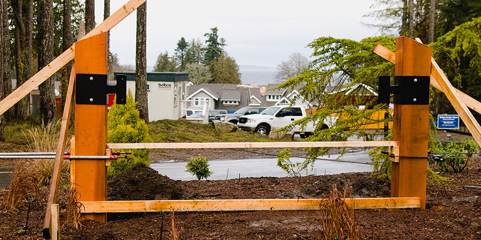 Construction site with wooden beams and support structures in place. Houses and a few vehicles are visible in the background among trees on a cloudy day.