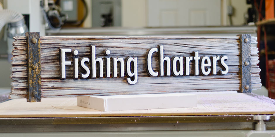 A wooden sign with the text "Fishing Charters" displayed prominently, set on a surface with blurred background elements.