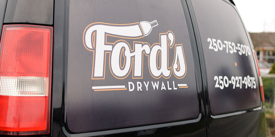 A black vehicle with "Ford's Drywall" and two phone numbers (250-752-5070 and 250-927-9675) displayed on the rear side window.