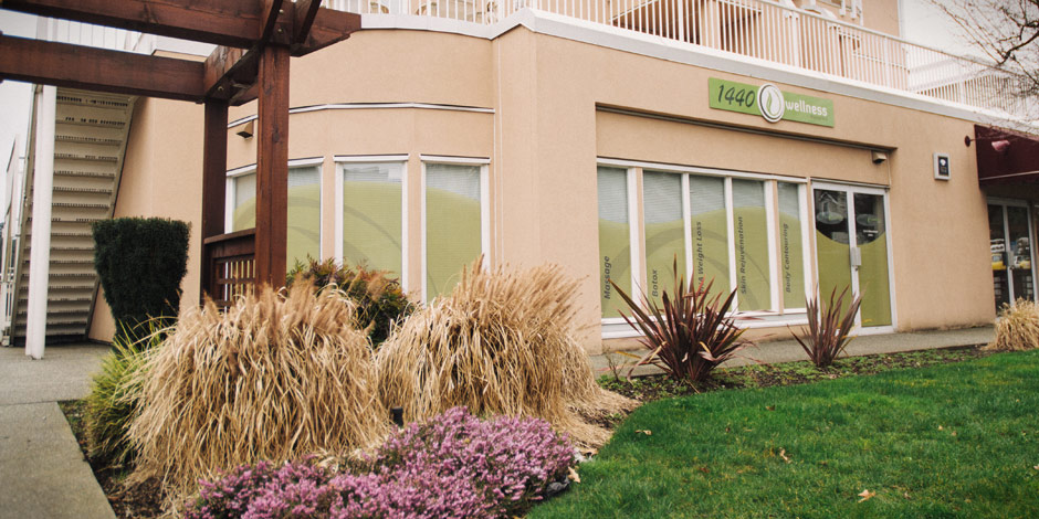A beige building with large windows, a "wellness" sign, and a green lawn with shrubs and plants in the foreground. An external staircase is visible on the left.