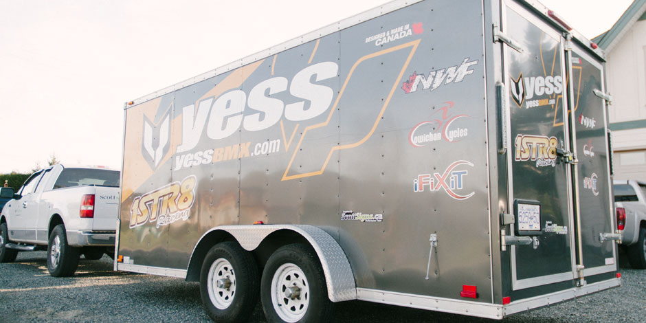 A silver trailer branded with “yess” and “yessbmx.com” is hitched to a white truck, with various decals and sponsors' logos visible on its side.