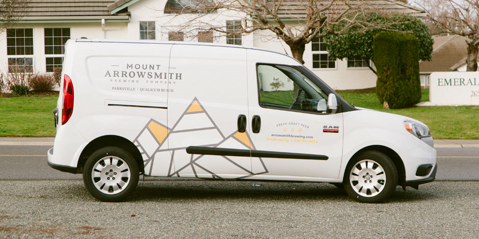 A white van labeled "Mount Arrowsmith Brewing Company" is parked on the street. The van has a geometric mountain design and contact information on its side. Houses and trees are in the background.