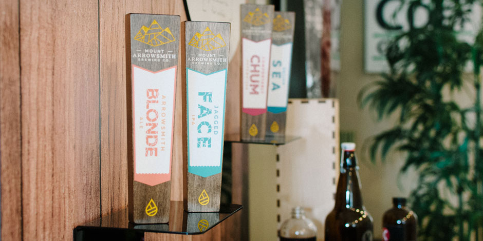 A display of Mount Arrowsmith Brewing Co. beer taps, including "Blonde" and "Jaded Face," with bottles on nearby shelves and a plant in the background.