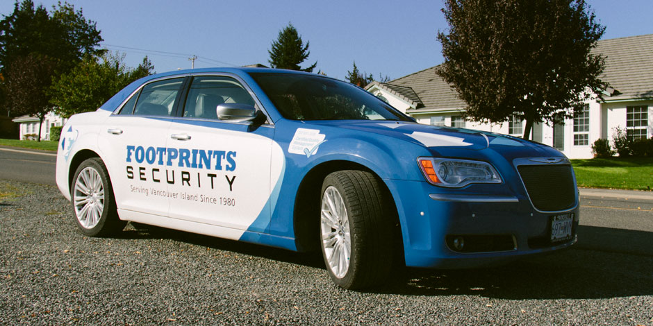 A blue and white Footprints Security patrol car is parked on a gravel area in front of a building. The text on the car reads "Serving Vancouver Island Since 1980".