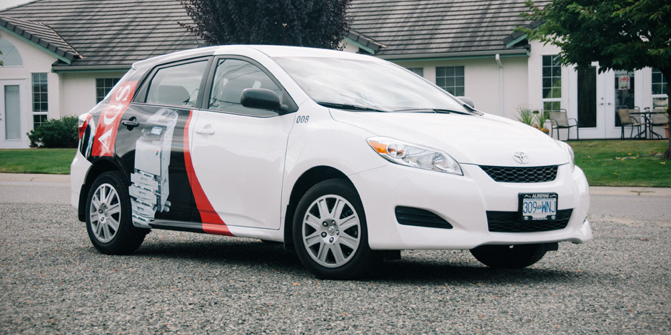 A white hatchback car with a graphic design on the side is parked on a gravel surface in front of a building with a grey roof.