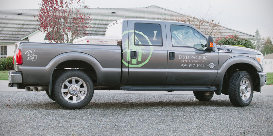 A gray pickup truck with company branding for "D&D Pacific Developments Ltd." featuring a phone number, parked on a gravel surface near a building.