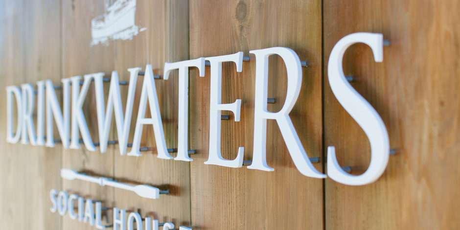 Close-up of a sign with white letters on a wooden background that reads "DRINKWATERS SOCIAL HOUSE." A paddle icon is visible below the text.