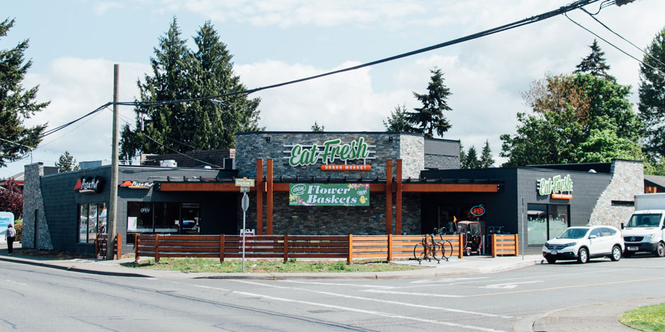 A street view of a corner grocery market named "Lift. Fresh Market" with a connected storefront "Flower Baskets" in a suburban area. Several cars and trees are visible in the background.