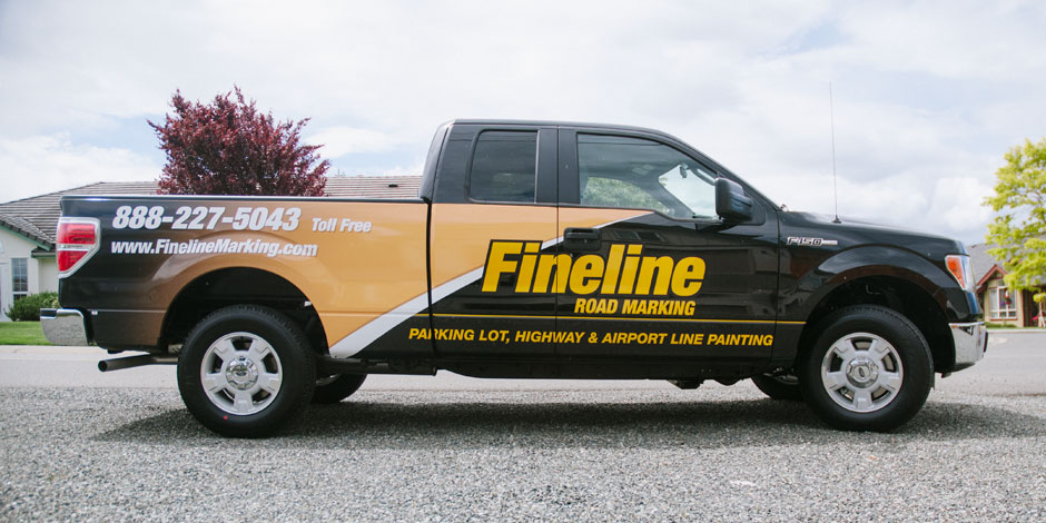 A black and yellow pick-up truck with "Fineline Road Marking" signage and contact information parked on a gravel driveway.