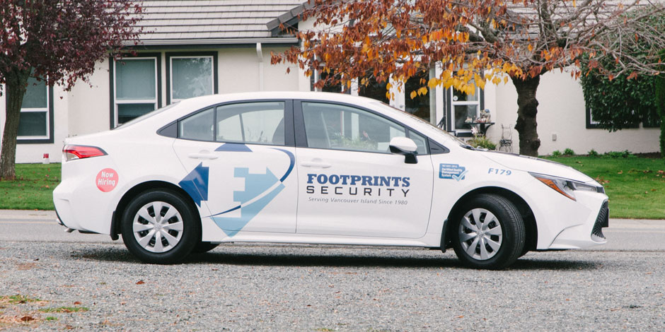 A white sedan with "Footprints Security" logo and text on its side is parked on a residential street.