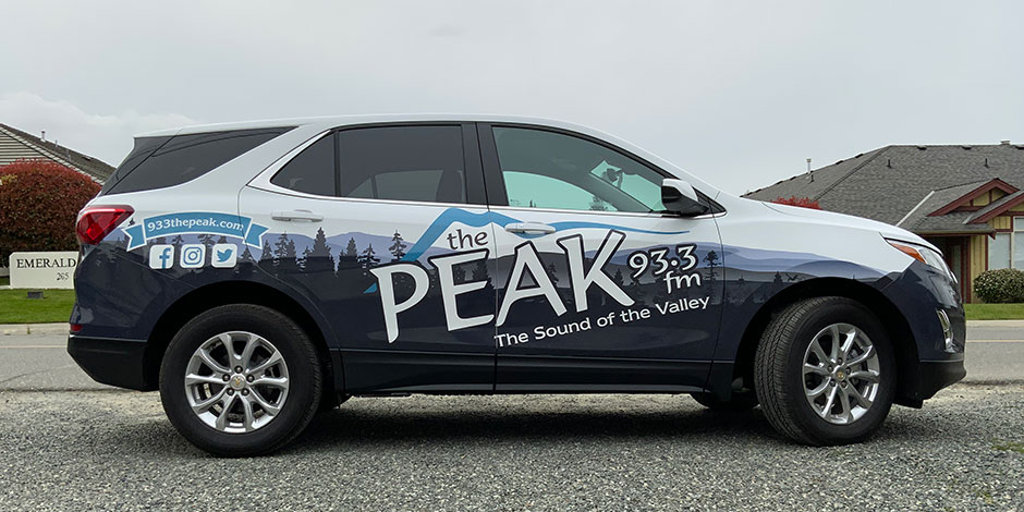 A branded SUV with "The Peak 93.3 FM" and "The Sound of the Valley" logos parked on a gravel surface in a suburban area. Social media icons and website URL are displayed on the vehicle.