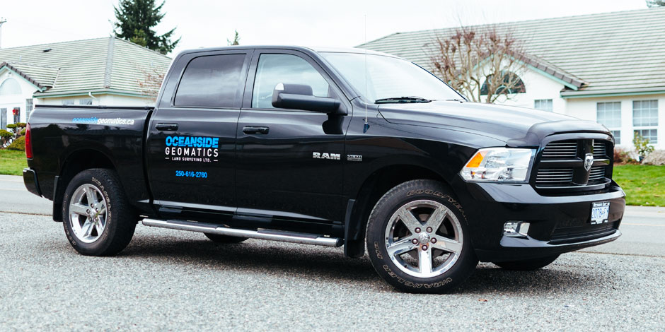 Black pickup truck with "Oceanside Geomatics" and contact information in blue text on the door, parked on a residential street with houses in the background.