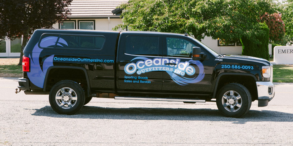 A black truck with "Oceanside Outfitters Inc" branding on the side, advertising sporting goods sales and rentals, parked on a street in front of a house and trees.