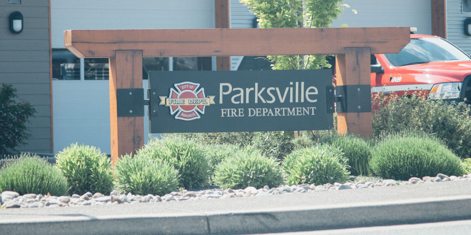 A sign for Parksville Fire Department stands in front of a building. A red emergency vehicle is partially visible in the background.