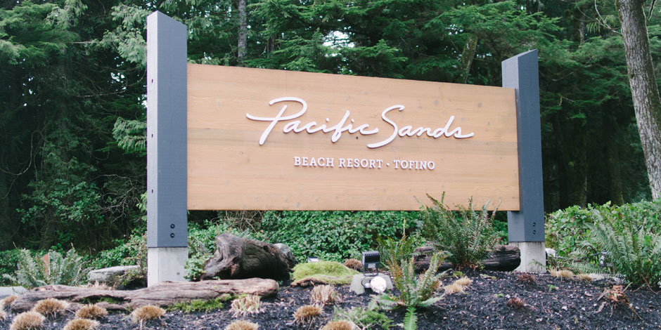 Sign for "Pacific Sands Beach Resort - Tofino" stands amidst landscaped greenery and tree-covered background.