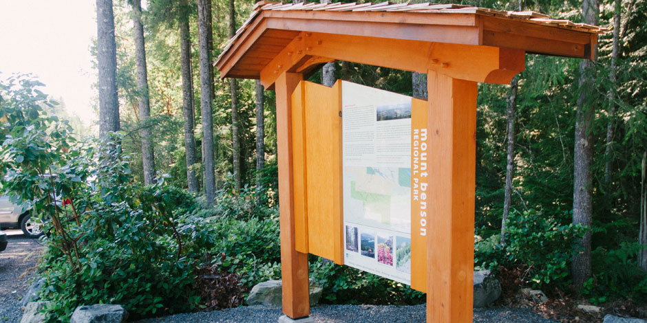 Wooden information kiosk in a forested area displaying a map and details about Mount Benson. Trees and parked cars are visible in the background.