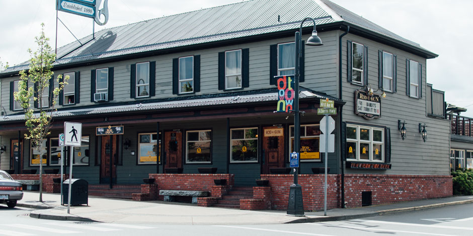 A two-story gray building with black trim and a metal roof. It has multiple windows, signs indicating "Black Bear Bread" and street signs in front of it. There is a pedestrian crossing nearby.