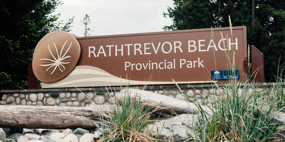 A brown and tan sign for Rathtrevor Beach Provincial Park is surrounded by driftwood and greenery, with trees visible in the background.