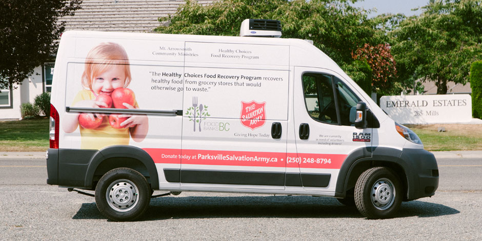 A white van with signage for the Healthy Choices Food Recovery Program, featuring a child holding apples, logos, and donation information. The van is parked on a residential street.