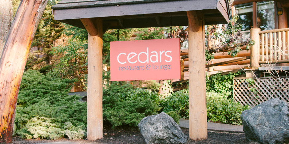 Signboard of Cedars Restaurant & Lounge hanging under a wooden archway, with a background of greenery and part of a log building.