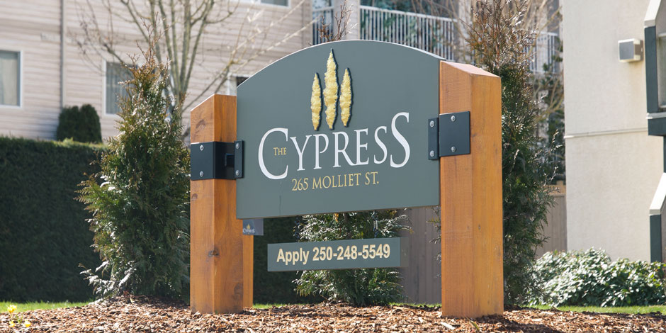 Sign with text "The Cypress, 265 Molliet St., Apply 250-248-5549" in front of a building with trees and shrubs in the background.