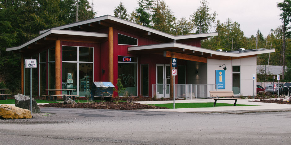 A modern building with a slanted roof, red and white exterior, and large windows serves as a visitor center. A sign indicates it is open. There are outdoor picnic tables and a parking area in the foreground.