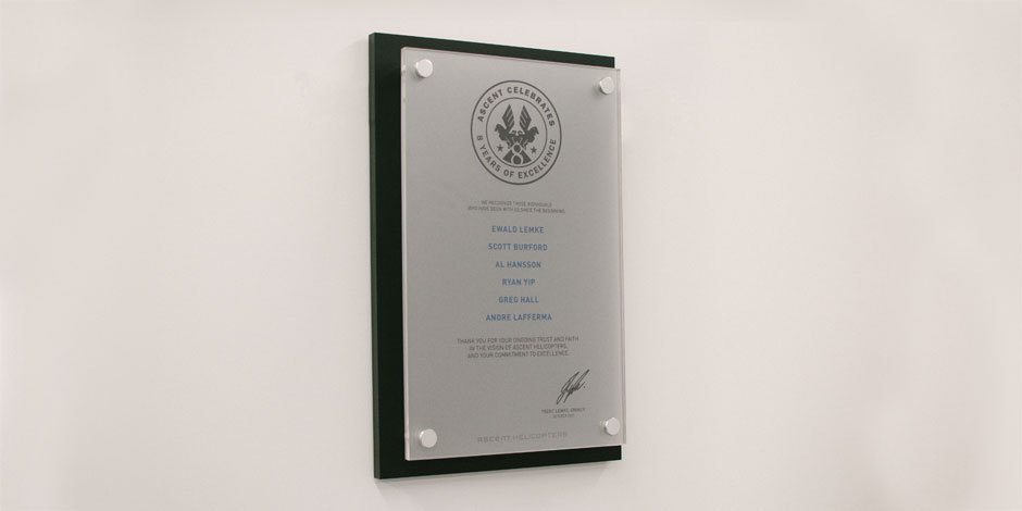 Plaque mounted on a wall recognizing individuals for their contributions. Top features an engraved emblem, and names listed below, with a signature at the bottom right.