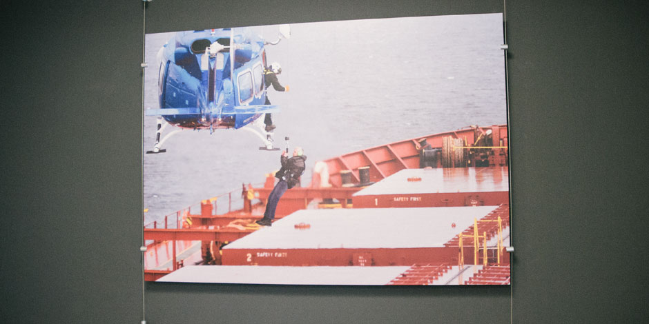 A photo hung on a wall depicts a person being hoisted from a red cargo ship by a blue helicopter hovering above.