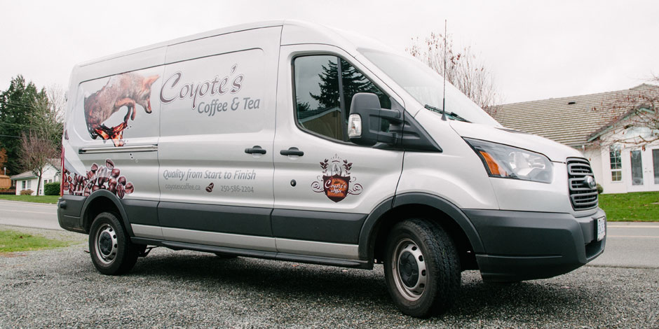 A white van with "Coyote's Coffee & Tea" branding is parked on a street. The van displays the company’s logo, contact information, and a graphic of a coyote on the side.