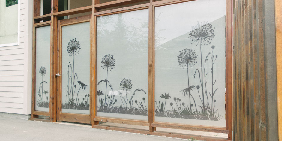 Large frosted windows with a floral pattern featuring dandelions, framed by wooden panels on a building exterior.