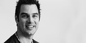 Black-and-white photo of a man with short, dark hair, smiling at the camera. He wears a casual shirt and a beaded necklace. The background is plain and light-colored.