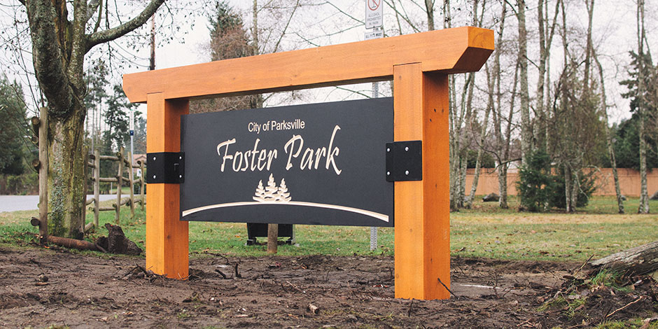 A wooden sign reading "City of Parksville Foster Park" stands in a grassy area with trees and a road in the background.