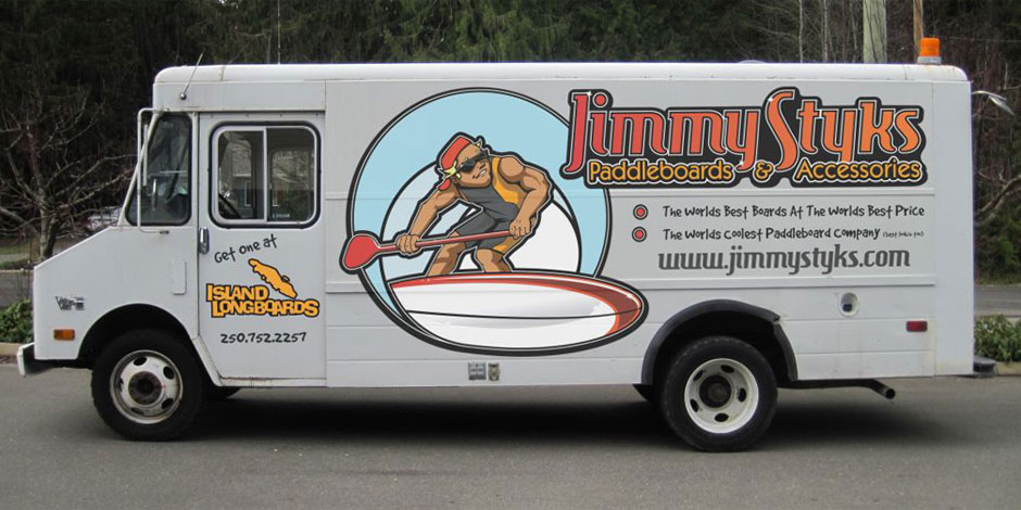 Side view of a white delivery truck with a large advertisement for Jimmy Styk's Paddleboards and Accessories. The ad shows a cartoon figure paddling a large board and includes contact details and website.