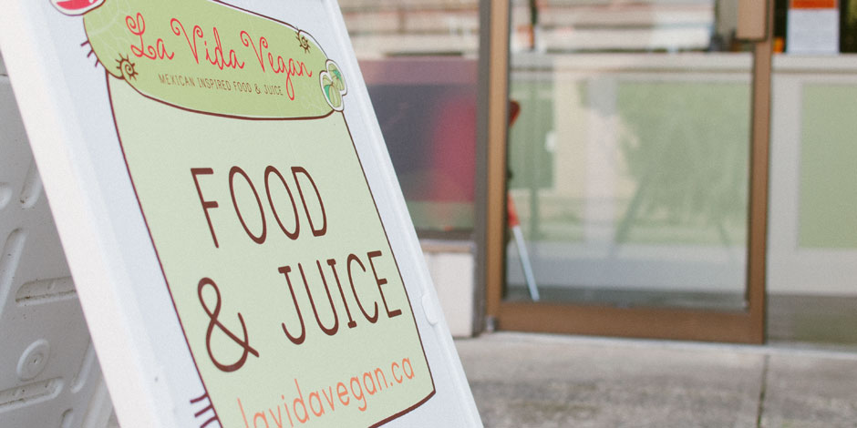 A sandwich board outside a shop displays a sign that reads, "La Vida Vegan - Mexican Inspired Food & Juice, FOOD & JUICE, lavidavegan.ca". The shop's entrance is visible in the background.