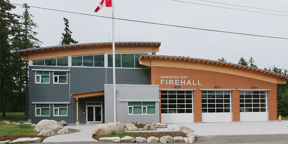 Front view of Nanoose Bay Firehall, showcasing a modern two-story building with large garage doors, a Canadian flag, and surrounded by trees.