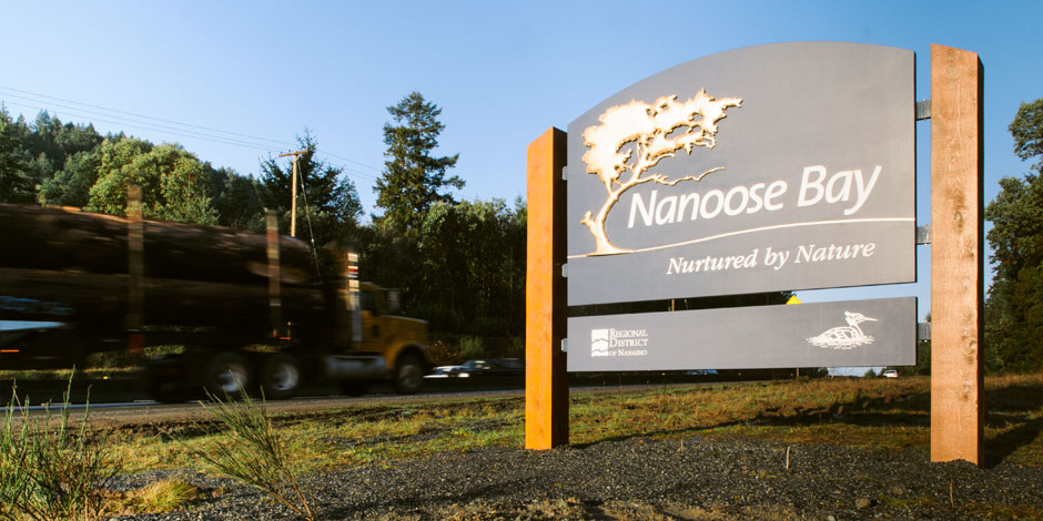 A sign reading "Nanoose Bay: Nurtured by Nature" stands by a roadside with a logging truck passing by and trees in the background.