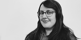 A picture of Naomi Cox with long hair and glasses, wearing a hoodie, looks at the camera with a slight smile, set against a plain background.