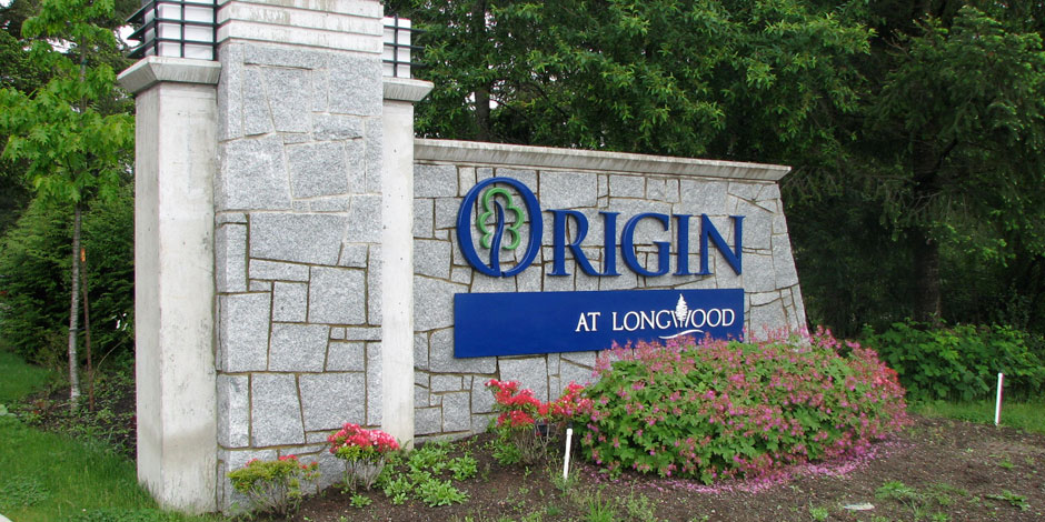 A stone monument sign with greenery reads "Origin at Longwood." The sign is set among landscaped shrubs and flowers.