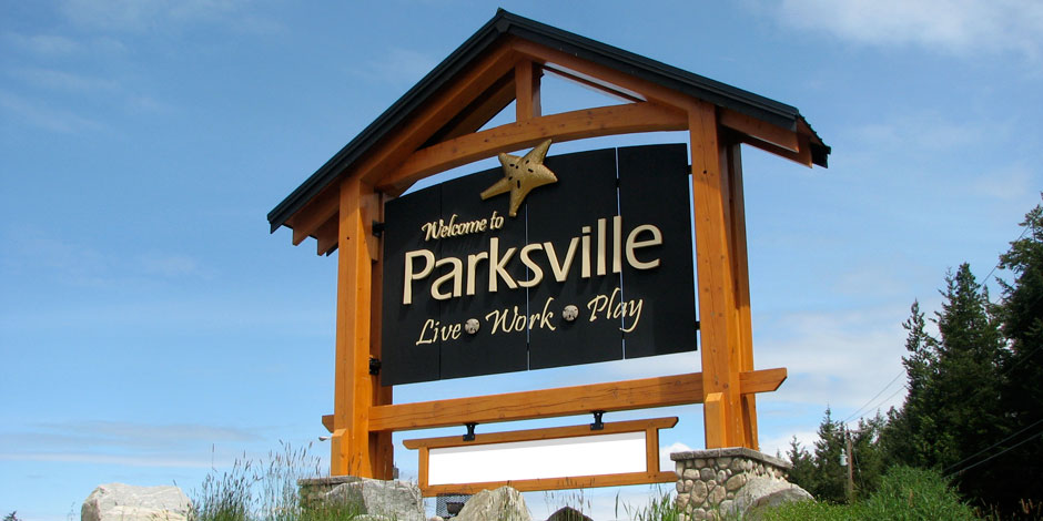 Wooden sign reading "Welcome to Parksville Live Work Play," with a star symbol at the top, set against a clear blue sky and greenery in the background.