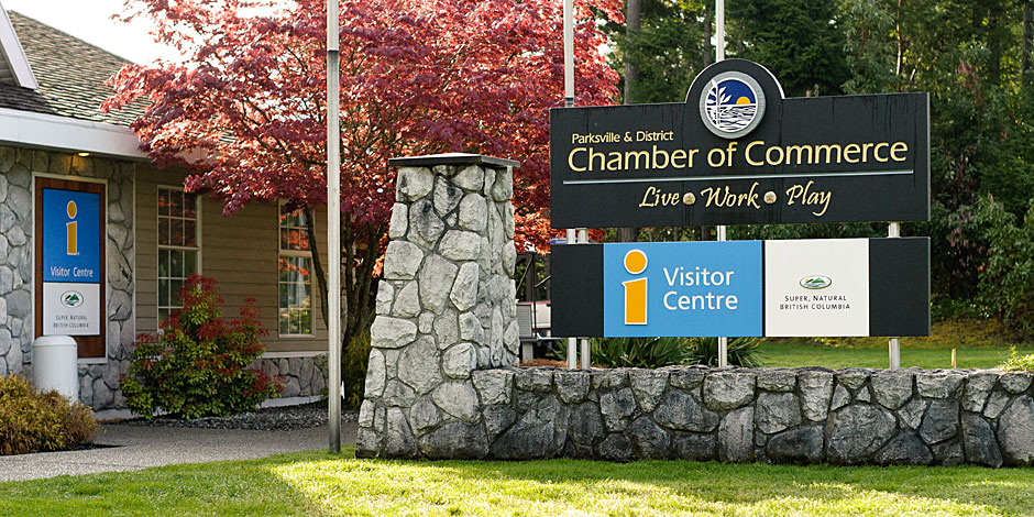 Image of the Parksville & District Chamber of Commerce and Visitor Centre building with signs displaying the name and logo, a nearby information sign, and landscaping including a tree and stone wall.