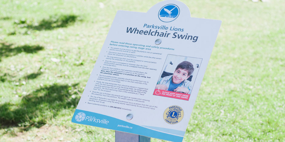 A signboard titled "Parksville Lions Wheelchair Swing" with user guidelines and safety procedures, placed in a grassy area. The board also features logos and an image of a smiling person.