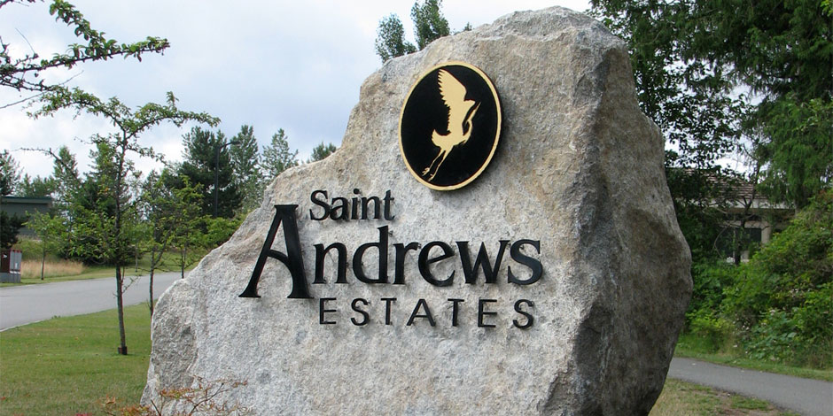 Sign for Saint Andrews Estates is engraved into a large rock, with a black and gold emblem of a bird at the top. Trees and a road are visible in the background.