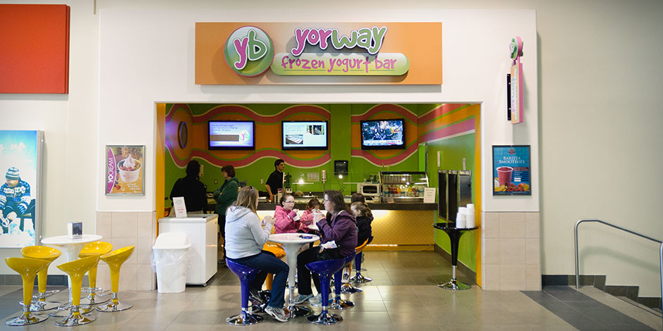 A frozen yogurt shop with colorful decor and signage. People are seated at vibrant tables enjoying their yogurt while others stand at the counter. Three advertisements are displayed on screens behind the counter.