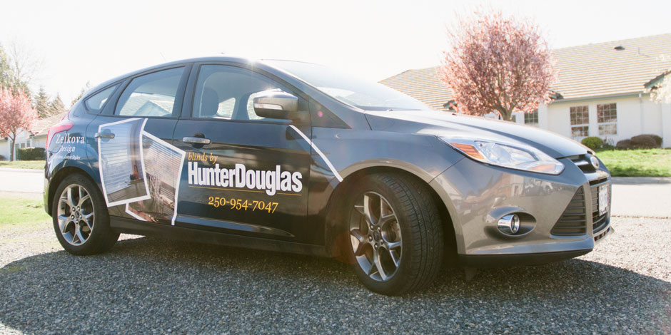 A gray car with "HunterDouglas" branding and a contact number, parked on a gravel driveway in front of a house and trees.