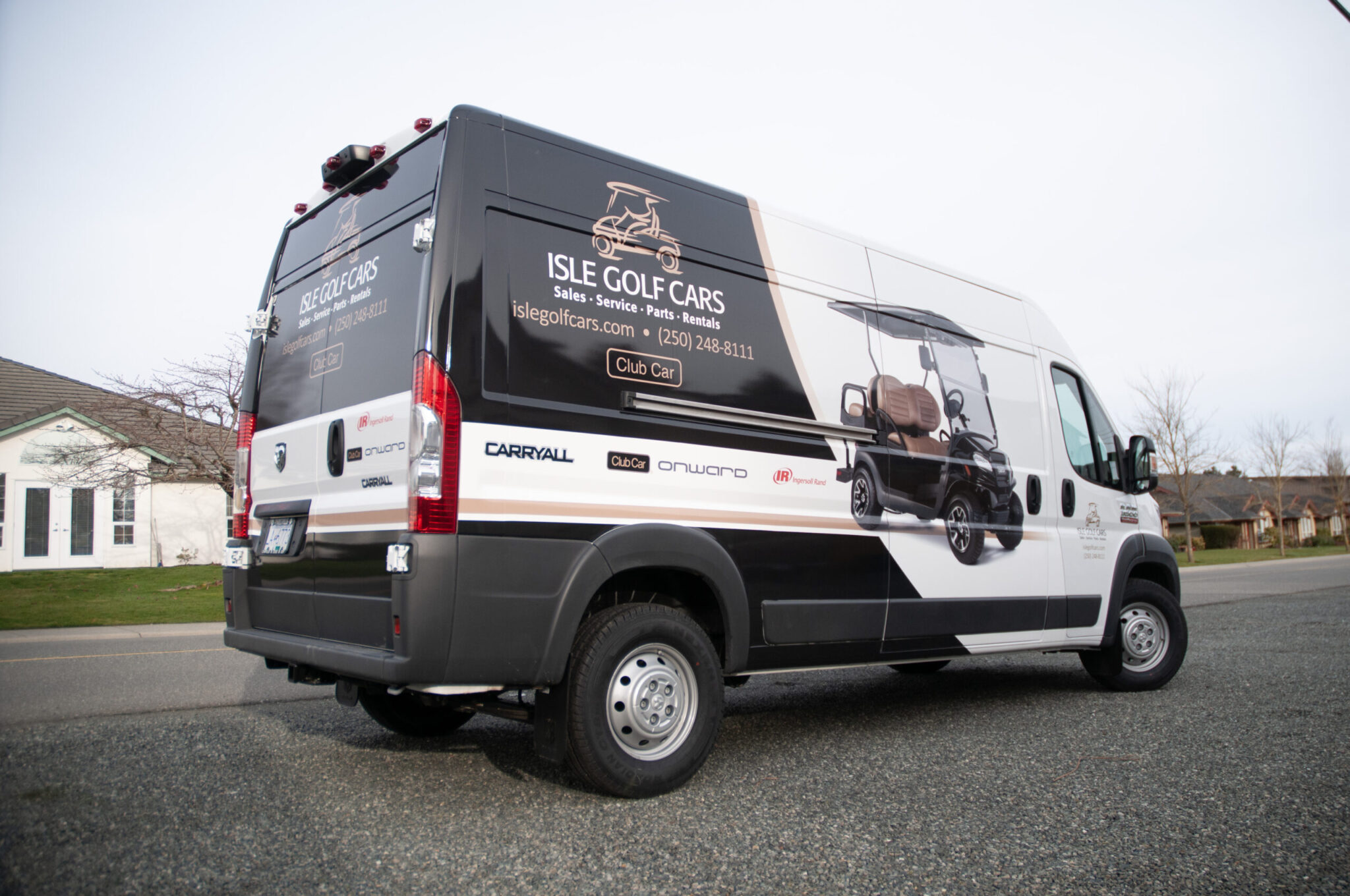 A white Isle Golf Cars service van is parked on a suburban street. The van has advertising graphics for sales, service, and rentals of golf carts, including contact details.