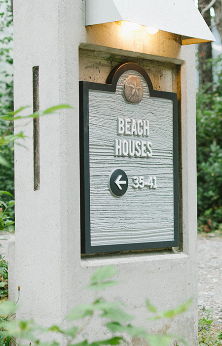 Sign reading “BEACH HOUSES 35-41” with an arrow pointing left, attached to a concrete structure and surrounded by greenery.