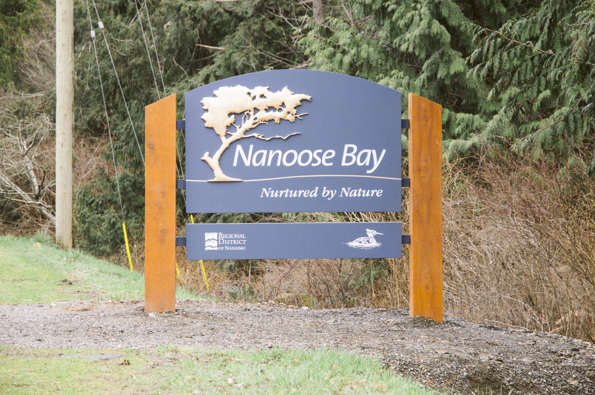 A sign for Nanoose Bay, reading "Nanoose Bay — Nurtured by Nature," stands with a depiction of a tree and bird, backed by forested vegetation.